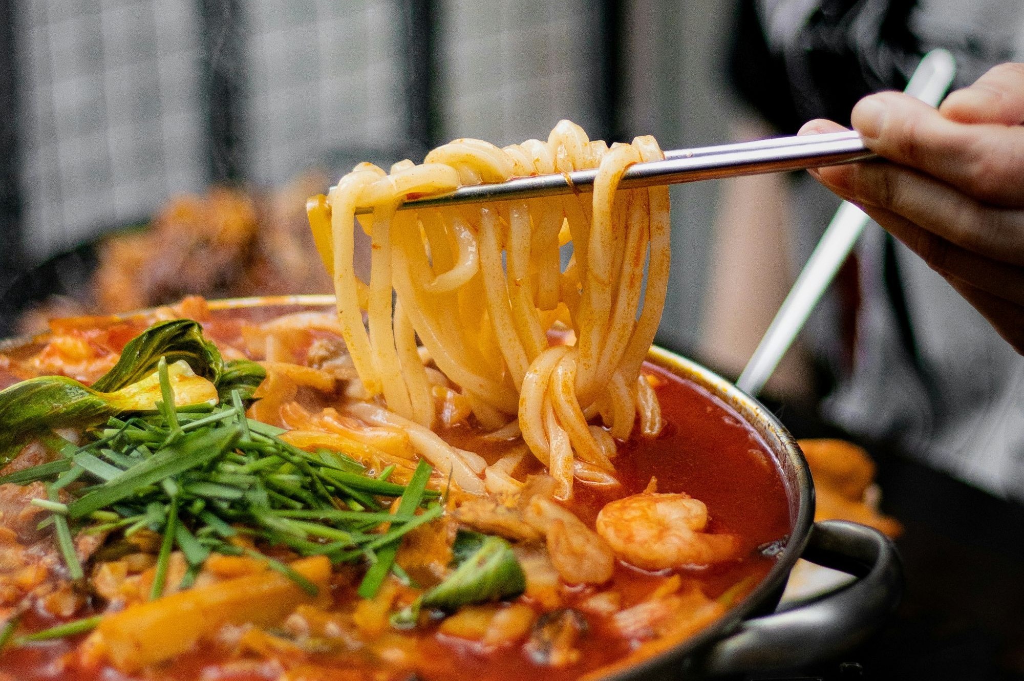 A person holding noodles with chopsticks above a spicy broth.