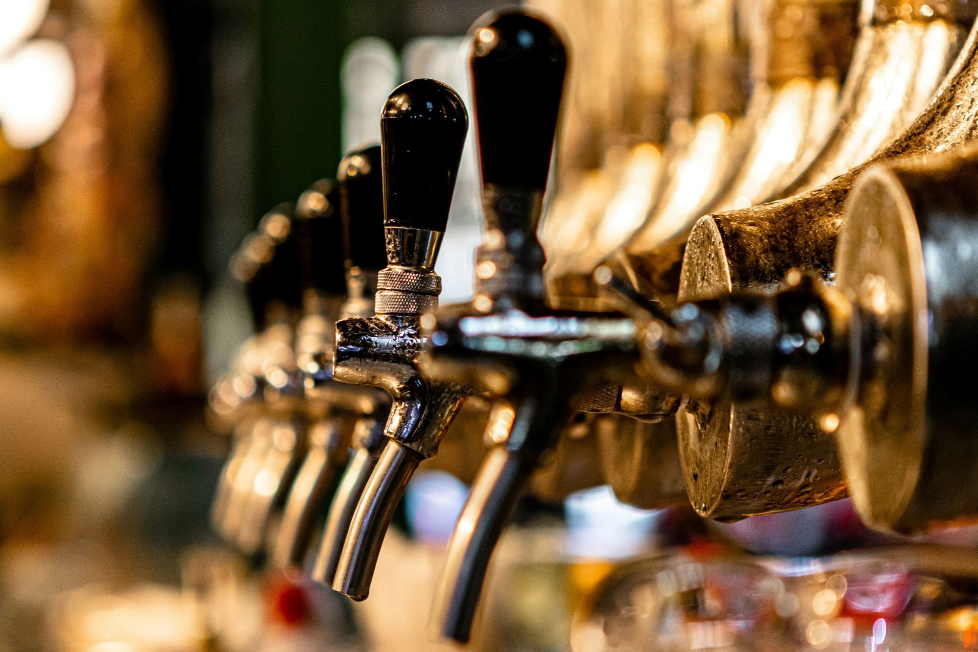 A close-up row of metallic draft beer taps with black handles at a dimly lit bar.