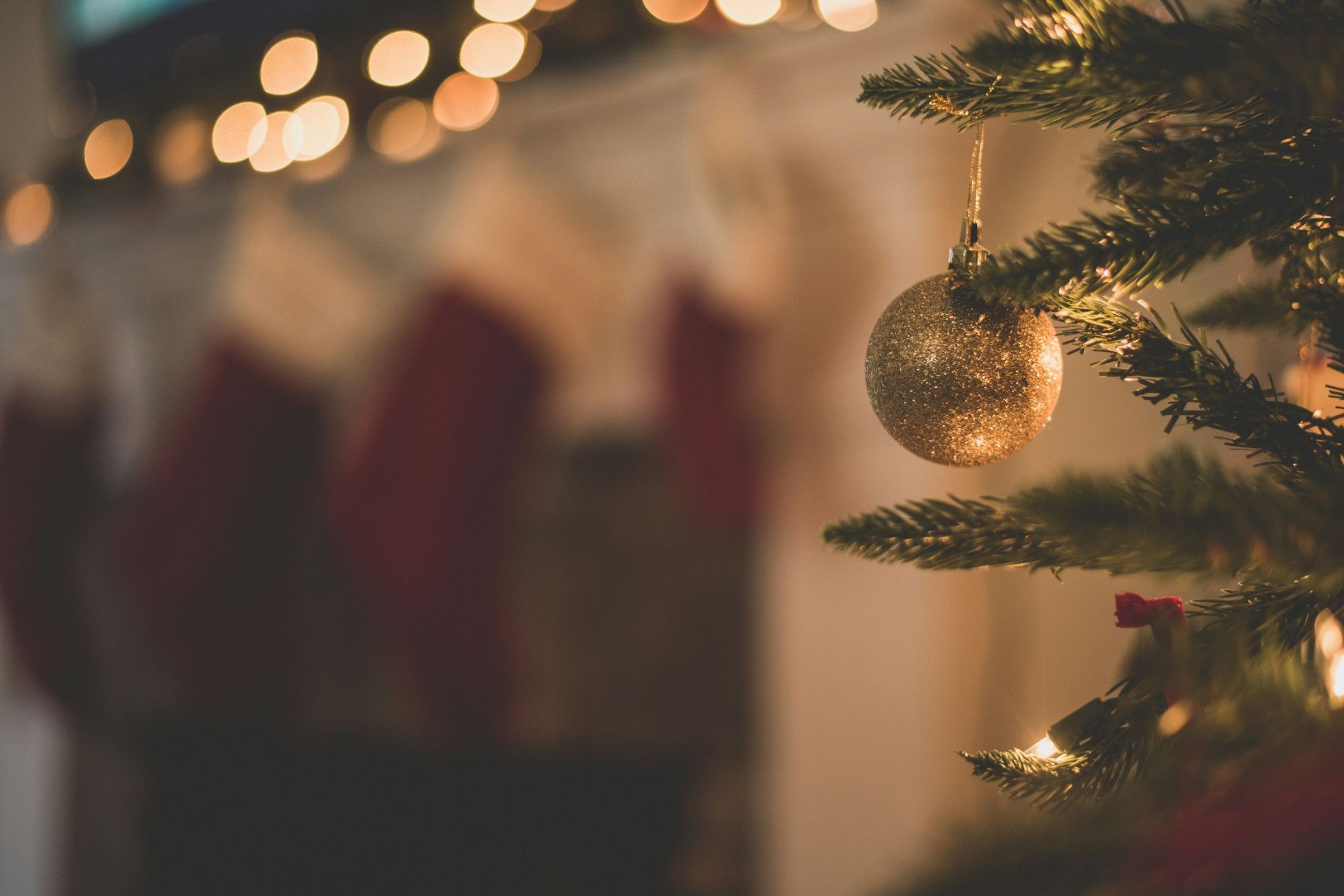 A golden bauble hanging from a Christmas tree with stockings in the background.