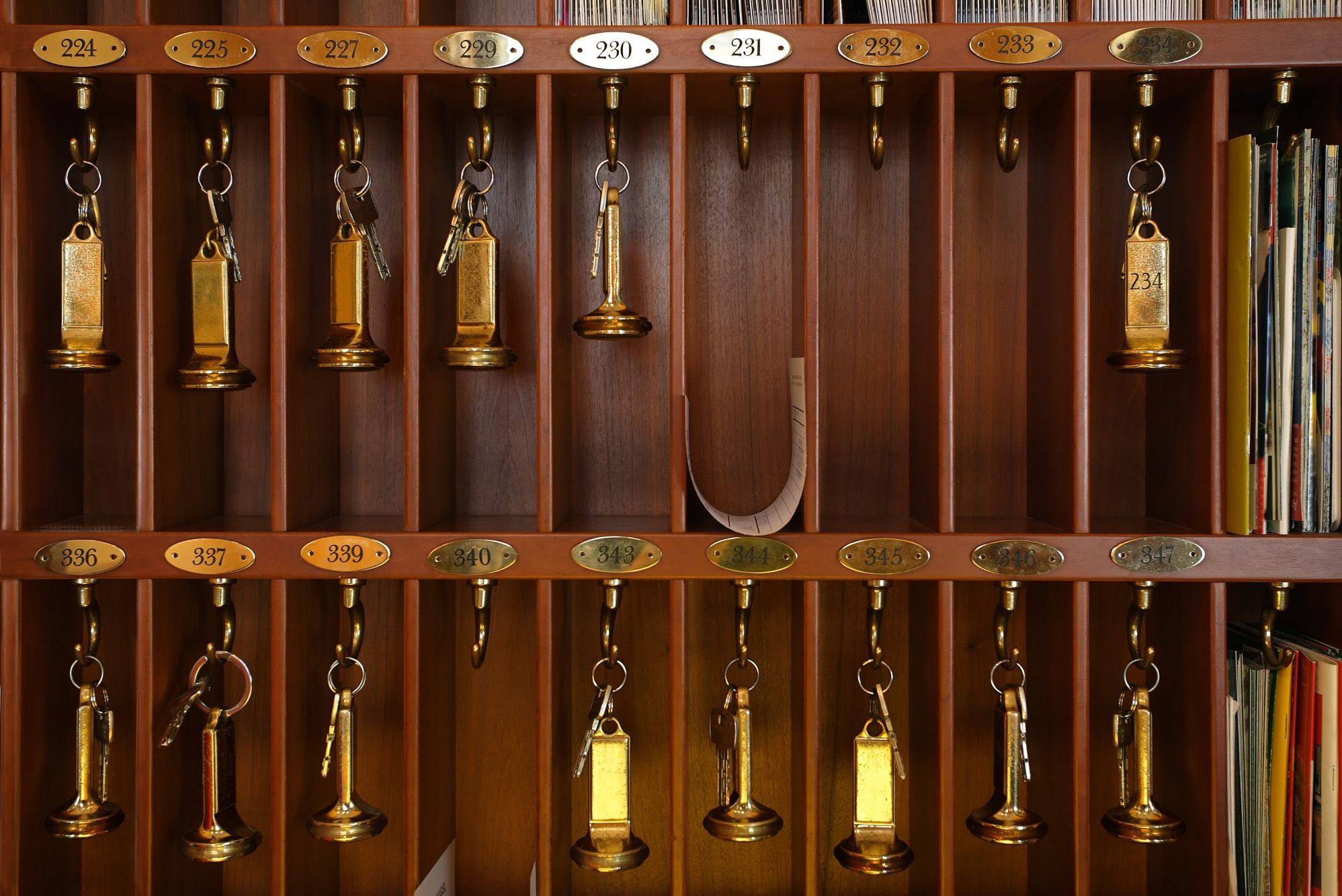  A traditional wooden hotel reception rack features two rows of numbered cubbies, most holding heavy brass keys on hooks while a few remain empty or contain papers.