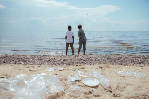Two children looking out across the sea with a pile of rubbish and pollution on the beach behind them.