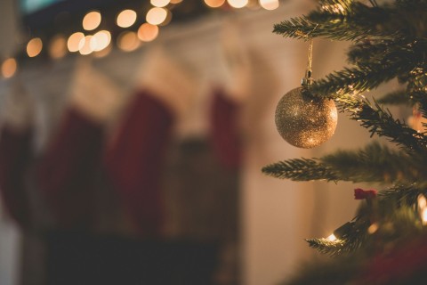 A golden bauble hanging from a Christmas tree with stockings in the background.