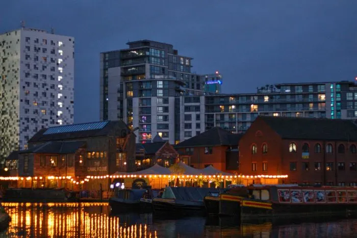 Narrowboats moored along a Birmingham canal at dusk, with warm outdoor lights reflecting in the water.