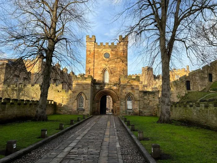 Durham Castle's stone gatehouse framed by two bare trees, with a cobbled pathway.