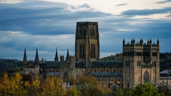 Durham Cathedral with its central tower and spires rising above autumn trees under a cloudy sky.