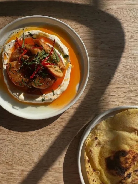 Two plates of food on a table with charred peaches, labneh, chilli, and flatbread.