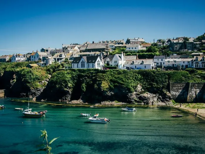A coastal village built along grassy cliffs overlooking a clear turquoise bay with several small boats floating on the water.