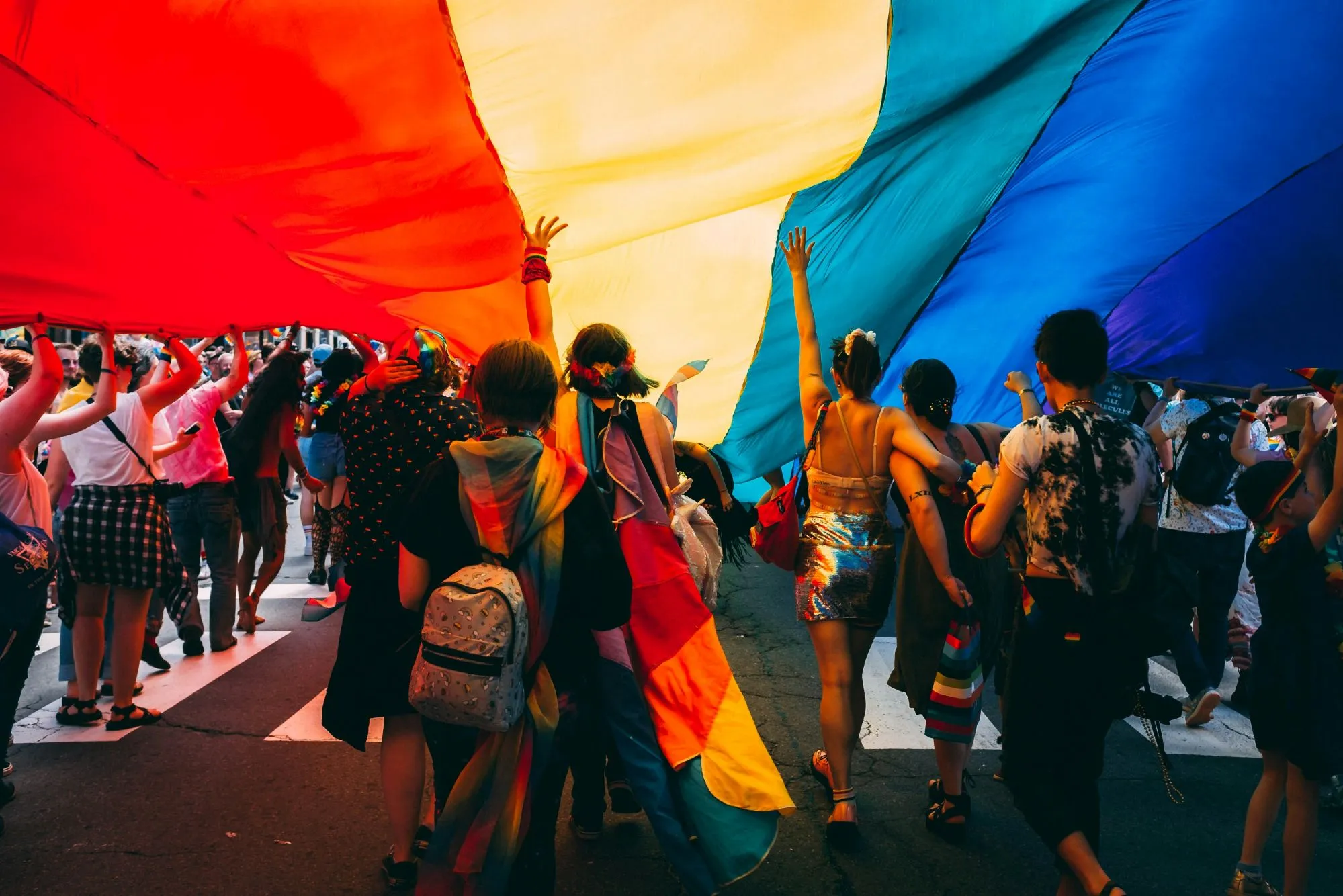 People marching in a Pride parade holding a rainbow flag above them.